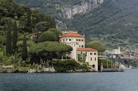 A picturesque villa is nestled on a lush hillside, surrounded by verdant gardens and sculpted trees. The elegant building features cream-colored walls and a red-tiled roof, sitting beside a tranquil body of water. The background reveals a mountainous landscape with scattered houses, emphasizing the secluded and serene location.