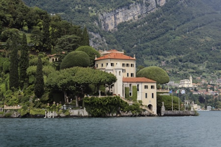 A picturesque villa is nestled on a lush hillside, surrounded by verdant gardens and sculpted trees. The elegant building features cream-colored walls and a red-tiled roof, sitting beside a tranquil body of water. The background reveals a mountainous landscape with scattered houses, emphasizing the secluded and serene location.