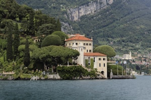 A picturesque villa is nestled on a lush hillside, surrounded by verdant gardens and sculpted trees. The elegant building features cream-colored walls and a red-tiled roof, sitting beside a tranquil body of water. The background reveals a mountainous landscape with scattered houses, emphasizing the secluded and serene location.