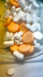 A variety of white and orange pills and tablets are scattered inside a container, with some featuring imprinted text or markings. The assortment includes both round and oblong shapes, suggesting different types or doses of medication.