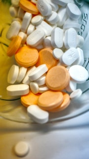 A variety of white and orange pills and tablets are scattered inside a container, with some featuring imprinted text or markings. The assortment includes both round and oblong shapes, suggesting different types or doses of medication.