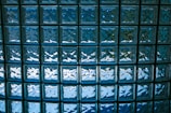 A grid of translucent glass blocks, each with a slightly convex surface, casting reflections and light patterns. The glass blocks are arranged in a uniform pattern, creating a mosaic-like effect with predominantly blue hues.