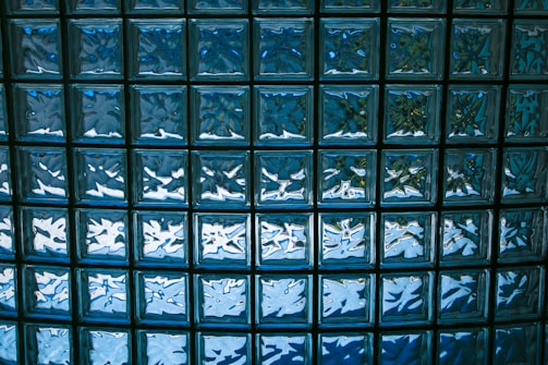 A grid of translucent glass blocks, each with a slightly convex surface, casting reflections and light patterns. The glass blocks are arranged in a uniform pattern, creating a mosaic-like effect with predominantly blue hues.
