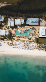 Aerial view of a beachside resort with modern buildings and a large swimming pool surrounded by palm trees. Rows of white lounge chairs and umbrellas line the sandy beach. The turquoise ocean stretches out at the bottom, gradually darkening in color. Dense greenery borders the top edge of the image.