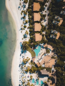 An aerial view of a coastal resort featuring several buildings with terracotta roofs surrounded by lush greenery. A large, uniquely-shaped swimming pool is at the center, bordered by sunbeds and shaded areas. The beach nearby showcases white sand curving along the shoreline, dotted with palm trees and beach chairs. The clear turquoise water of the sea contrasts with the white sand and the greenery.