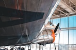 A close-up view of a vintage airplane's undercarriage, with shiny metallic surfaces and a visible propeller. The aircraft is situated inside a hangar with large windows allowing natural light to illuminate the scene. Another aircraft can be seen in the background, partially visible, adding to the setting's historical and aeronautical theme.