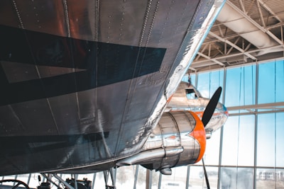 A close-up view of a vintage airplane's undercarriage, with shiny metallic surfaces and a visible propeller. The aircraft is situated inside a hangar with large windows allowing natural light to illuminate the scene. Another aircraft can be seen in the background, partially visible, adding to the setting's historical and aeronautical theme.