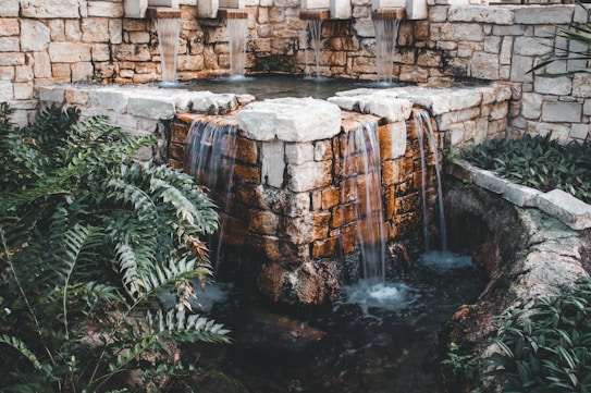 A stone fountain with multiple streams of water cascading down into a small pool bordered by lush, green foliage. The walls of the fountain are constructed from weathered stones, giving it a rustic appearance, and the surrounding plants add a touch of natural beauty.