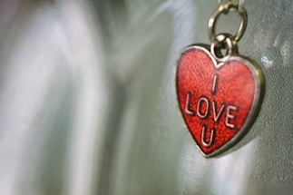 A heart-shaped keychain with a red enamel surface and the words 'I LOVE U' embossed on it, hanging against a blurred background.