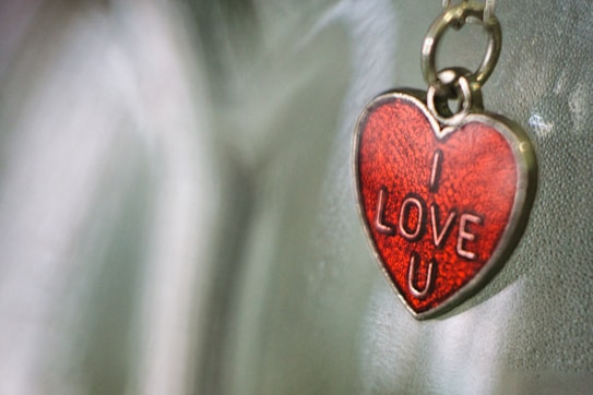 A heart-shaped keychain with a red enamel surface and the words 'I LOVE U' embossed on it, hanging against a blurred background.