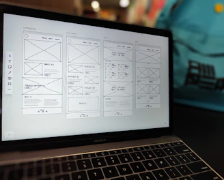 A laptop displaying a wireframe design for a website. The screen shows multiple pages with sections labeled for a landing page, product details, and about information. The background contains a blurred backpack.