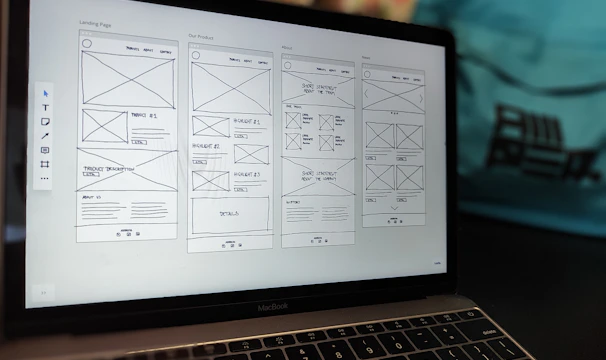 A laptop displaying a wireframe design for a website. The screen shows multiple pages with sections labeled for a landing page, product details, and about information. The background contains a blurred backpack.