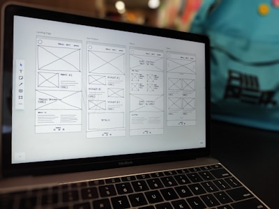 A laptop displaying a wireframe design for a website. The screen shows multiple pages with sections labeled for a landing page, product details, and about information. The background contains a blurred backpack.