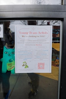A poster on a window is calling for submissions from young trans artists to design an LGBTQ+ pride themed mascot. The poster details the contest, offering a cash prize and the chance to have artwork displayed. It includes a QR code for more information and features a small depiction of a green dragon holding a rainbow flag. A blue plush toy with 'Gifts for Humans' written on it is visible next to the poster.