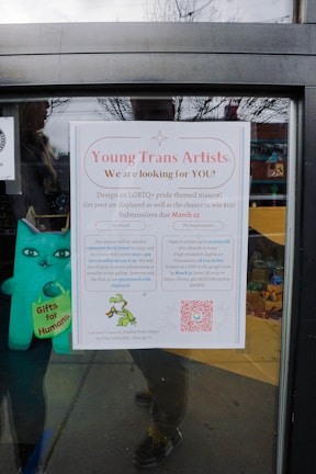 A poster on a window is calling for submissions from young trans artists to design an LGBTQ+ pride themed mascot. The poster details the contest, offering a cash prize and the chance to have artwork displayed. It includes a QR code for more information and features a small depiction of a green dragon holding a rainbow flag. A blue plush toy with 'Gifts for Humans' written on it is visible next to the poster.