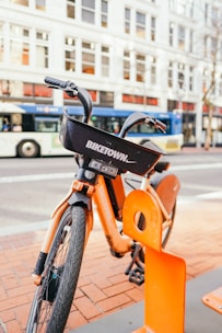 An orange rental bike is parked at a docking station on a city street. The bike displays a 'BIKETOWN' logo on its basket, and a large bus is visible in the background along with a row of buildings with numerous windows.