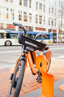 An orange rental bike is parked at a docking station on a city street. The bike displays a 'BIKETOWN' logo on its basket, and a large bus is visible in the background along with a row of buildings with numerous windows.