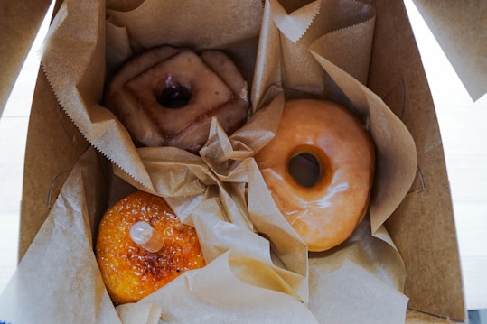 Three donuts are displayed inside a cardboard box with brown paper lining. The donuts include a traditional glazed donut, a square-shaped donut with a glaze on top, and a br&ucirc;l&eacute;e-style donut with a caramelized sugar top and a small pipette filled with a clear liquid.