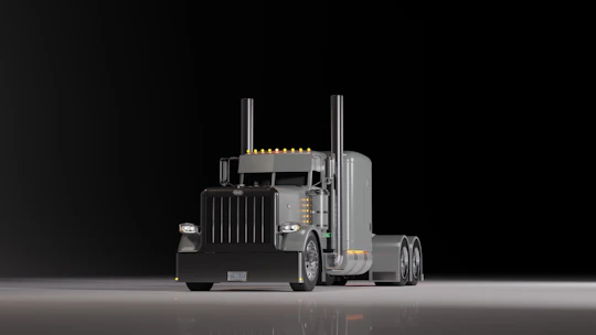 A large, shiny semi-truck with a prominent front grille is positioned on a reflective surface against a dark background. The truck features dual exhaust stacks and a sleeper cab design with several orange lights along the roofline.