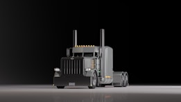 A large, shiny semi-truck with a prominent front grille is positioned on a reflective surface against a dark background. The truck features dual exhaust stacks and a sleeper cab design with several orange lights along the roofline.