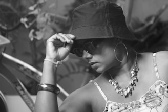 A person wearing a bucket hat, sunglasses, and hoop earrings, accessorized with a beaded necklace. The individual appears thoughtful, with one hand adjusting the hat. The background includes a bicycle and some blurred plants, all in black and white.