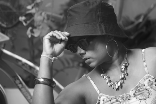 A person wearing a bucket hat, sunglasses, and hoop earrings, accessorized with a beaded necklace. The individual appears thoughtful, with one hand adjusting the hat. The background includes a bicycle and some blurred plants, all in black and white.