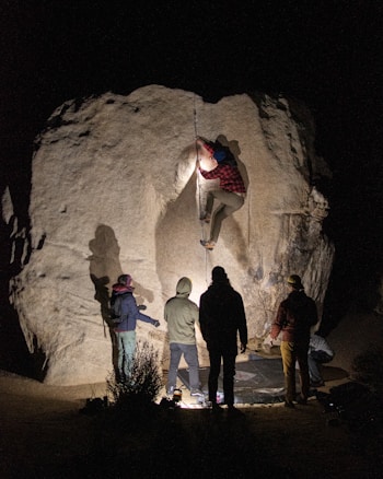 A climber is ascending a large rock formation at night, surrounded by supportive people on the ground. They are equipped with climbing gear and illuminated by artificial lights, creating dramatic shadows.