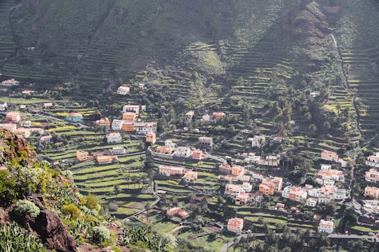 A picturesque village nestled in a lush, mountainous landscape with terraced fields. Numerous houses with orange and white facades are scattered across the hillside, surrounded by verdant greenery. The terrain features layered farming plots and a variety of trees dotting the slopes.
