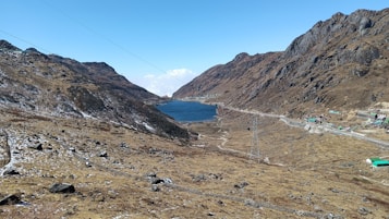 A stunning mountain landscape features a serene blue lake nestled between rocky hills. The terrain is rugged, with brown and green hues dominating the slopes. A road winds alongside the lake, leading to small buildings with green roofs scattered throughout the scene. The sky is clear and blue, enhancing the crisp, natural beauty of the setting.
