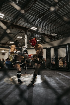 Two athletes are engaged in a mixed martial arts (MMA) fight inside a caged ring. Both fighters wear protective headgear and other gear, indicative of a training or sparring session. The environment is a large indoor gym with an audience observing the match through the cage.