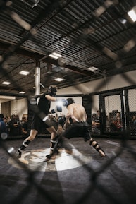 Two fighters engage in a mixed martial arts match within a caged ring. They are wearing protective gear, including gloves and shin guards, and are surrounded by spectators watching intently. Harsh lighting from above casts shadows on the fighters and the mat.