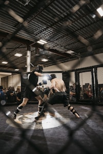 Two fighters engage in a mixed martial arts match within a caged ring. They are wearing protective gear, including gloves and shin guards, and are surrounded by spectators watching intently. Harsh lighting from above casts shadows on the fighters and the mat.