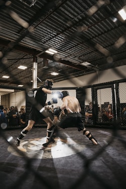 Two fighters engage in a mixed martial arts match within a caged ring. They are wearing protective gear, including gloves and shin guards, and are surrounded by spectators watching intently. Harsh lighting from above casts shadows on the fighters and the mat.