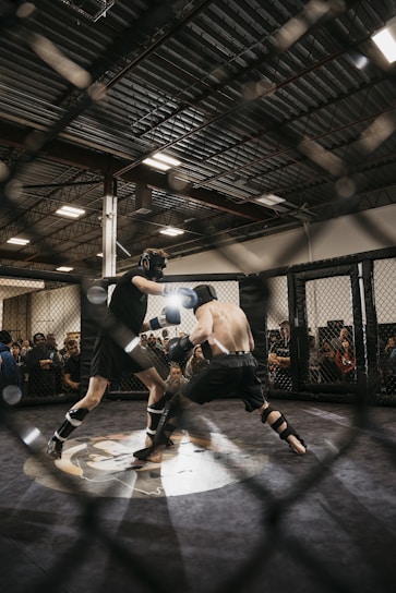 Two fighters engage in a mixed martial arts match within a caged ring. They are wearing protective gear, including gloves and shin guards, and are surrounded by spectators watching intently. Harsh lighting from above casts shadows on the fighters and the mat.