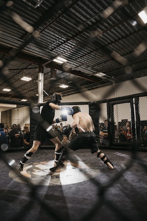 Two fighters engage in a mixed martial arts match within a caged ring. They are wearing protective gear, including gloves and shin guards, and are surrounded by spectators watching intently. Harsh lighting from above casts shadows on the fighters and the mat.