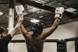 A person wearing a black helmet and white boxing gloves raises their arms in victory. An individual beside them, with a tattooed arm, is holding up one of their hands. The background features an indoor arena with a metal roof and lighting fixtures.