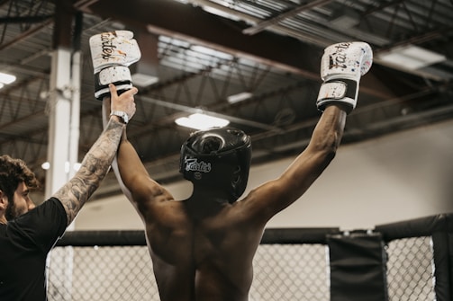 A person wearing a black helmet and white boxing gloves raises their arms in victory. An individual beside them, with a tattooed arm, is holding up one of their hands. The background features an indoor arena with a metal roof and lighting fixtures.