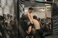 Two fighters engage in a mixed martial arts match inside a fenced cage, surrounded by audience members who are watching intently. The fighters are in close proximity, with one attempting a grapple move against the cage.