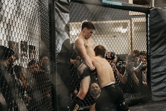 Two fighters engage in a mixed martial arts match inside a fenced cage, surrounded by audience members who are watching intently. The fighters are in close proximity, with one attempting a grapple move against the cage.