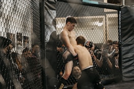 Two fighters engage in a mixed martial arts match inside a fenced cage, surrounded by audience members who are watching intently. The fighters are in close proximity, with one attempting a grapple move against the cage.