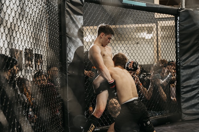 Two fighters engage in a mixed martial arts match inside a fenced cage, surrounded by audience members who are watching intently. The fighters are in close proximity, with one attempting a grapple move against the cage.