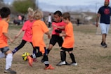 A group of children is playing soccer on a grassy field. They are dressed in bright orange and pink jerseys, focusing on controlling the ball. A coach or parent stands in the background, observing the game. The scene takes place outdoors with a clear sky and some trees in the distance.