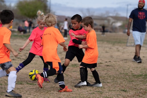 A group of children is playing soccer on a grassy field. They are dressed in bright orange and pink jerseys, focusing on controlling the ball. A coach or parent stands in the background, observing the game. The scene takes place outdoors with a clear sky and some trees in the distance.