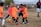 A group of children is playing soccer on a grassy field. They are dressed in bright orange and pink jerseys, focusing on controlling the ball. A coach or parent stands in the background, observing the game. The scene takes place outdoors with a clear sky and some trees in the distance.