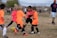 A group of children is playing soccer on a grassy field. They are dressed in bright orange and pink jerseys, focusing on controlling the ball. A coach or parent stands in the background, observing the game. The scene takes place outdoors with a clear sky and some trees in the distance.