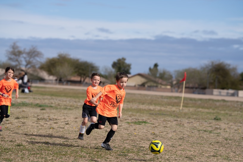 Children are playing soccer on a grassy field. The foreground shows three kids wearing orange jerseys running and chasing a soccer ball. The background showcases a rural setting with houses, trees, and a partly cloudy sky.