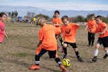 A group of young children playing soccer on a grassy field. They are wearing bright orange jerseys with black shorts, focusing intently on the game as they chase a soccer ball. The sky is partly cloudy, and there are trees and other players visible in the background.