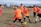 A group of young children playing soccer on a grassy field. They are wearing bright orange jerseys with black shorts, focusing intently on the game as they chase a soccer ball. The sky is partly cloudy, and there are trees and other players visible in the background.