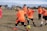 A group of young children playing soccer on a grassy field. They are wearing bright orange jerseys with black shorts, focusing intently on the game as they chase a soccer ball. The sky is partly cloudy, and there are trees and other players visible in the background.