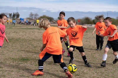 A group of young children playing soccer on a grassy field. They are wearing bright orange jerseys with black shorts, focusing intently on the game as they chase a soccer ball. The sky is partly cloudy, and there are trees and other players visible in the background.